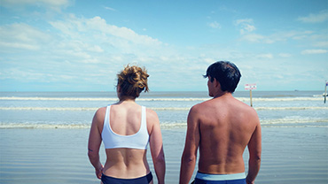 Two people in swimsuits standing on the beach looking out at the ocean and waves. Credit: NOAA